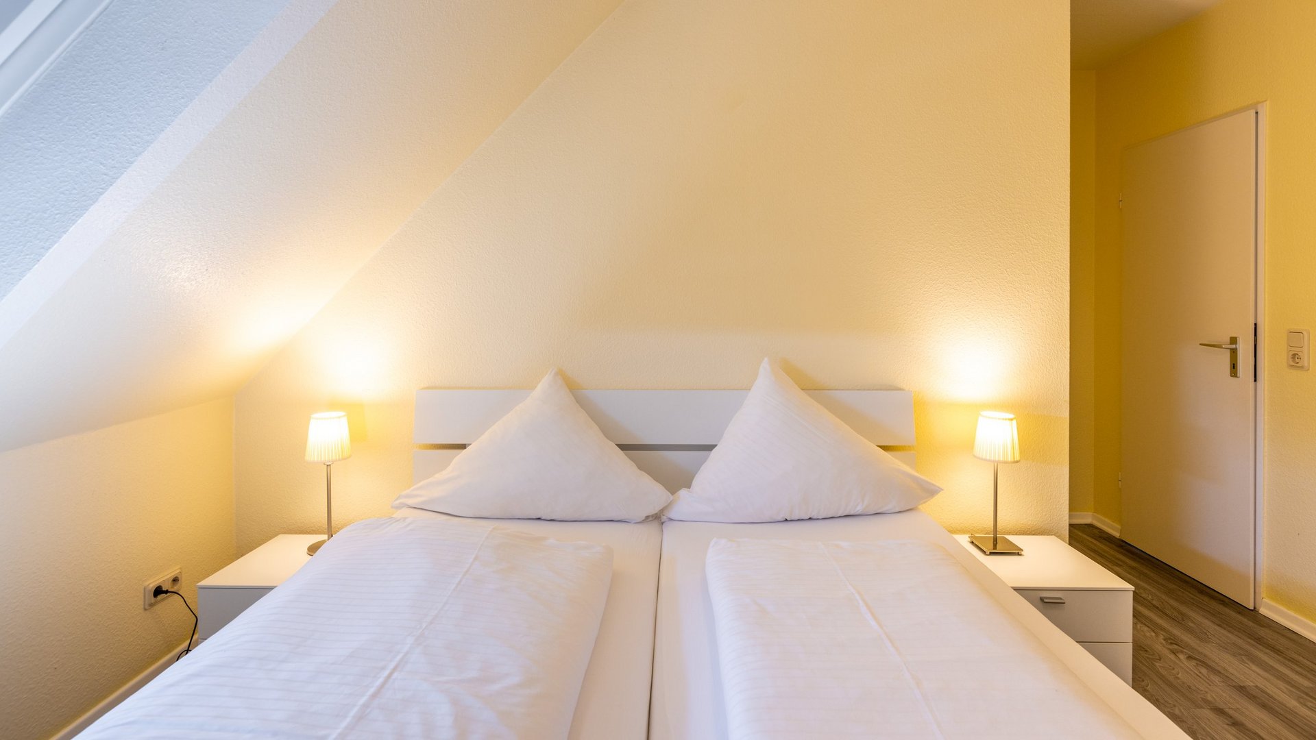 Gemünder Ferienpark: Jobs, die erfüllen Gemütliches Schlafzimmer mit Doppelbett, weißen Kissen und zwei Nachttischlampen