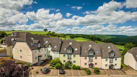 Ferien in der Eifel: ein erster Einblick Wohnhäuser vor grünen Hügeln unter bewölktem blauen Himmel