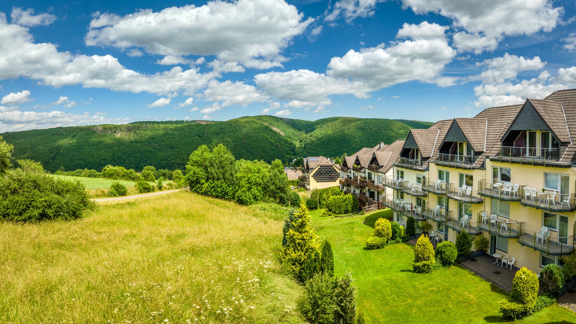So erreichen Sie unsere Ferienwohnungen in Gemünd. Gebäude am grünen Hügel mit bewölktem Himmel und Bergen im Hintergrund