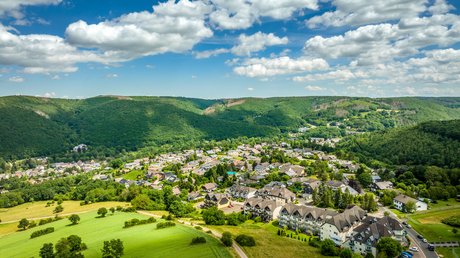 Ferien in der Eifel: ein erster Einblick Luftaufnahme eines Dorfes umgeben von grünen Wäldern und Feldern unter blauem Himmel