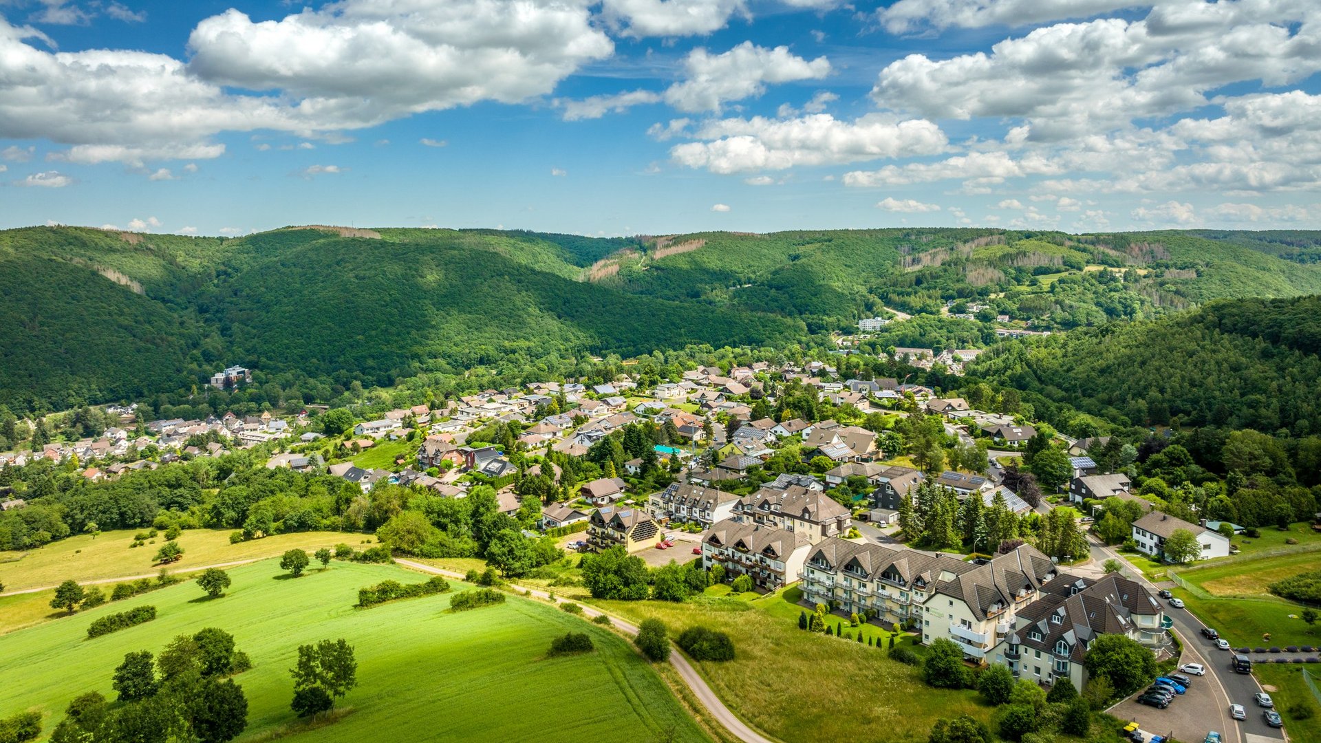 Wohnung mieten in Gemünd? Da haben wir was für Sie! Luftaufnahme eines Dorfes umgeben von grünen Wäldern und Feldern unter blauem Himmel