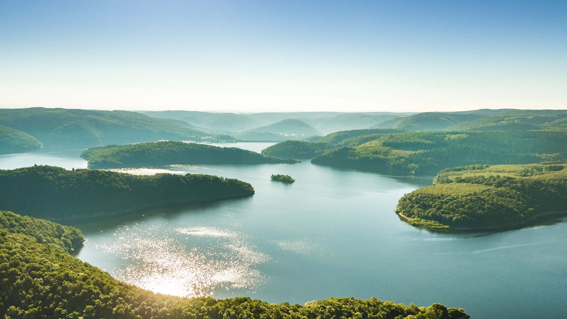 So erreichen Sie unsere Ferienwohnungen in Gemünd. Luftaufnahme eines großen, von Wald bedeckten Stausees mit leuchtendem Wasser