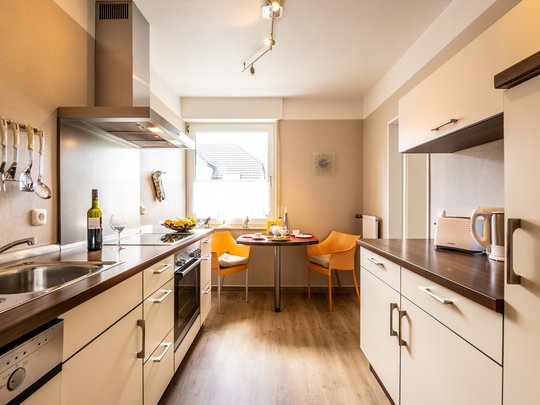 Ihre Unterkunft in Gemünd – Langzeitaufenthalt mit extra Service Moderne Küche mit Esstisch, zwei orangenen Stühlen, Weinflasche und Küchengeräten