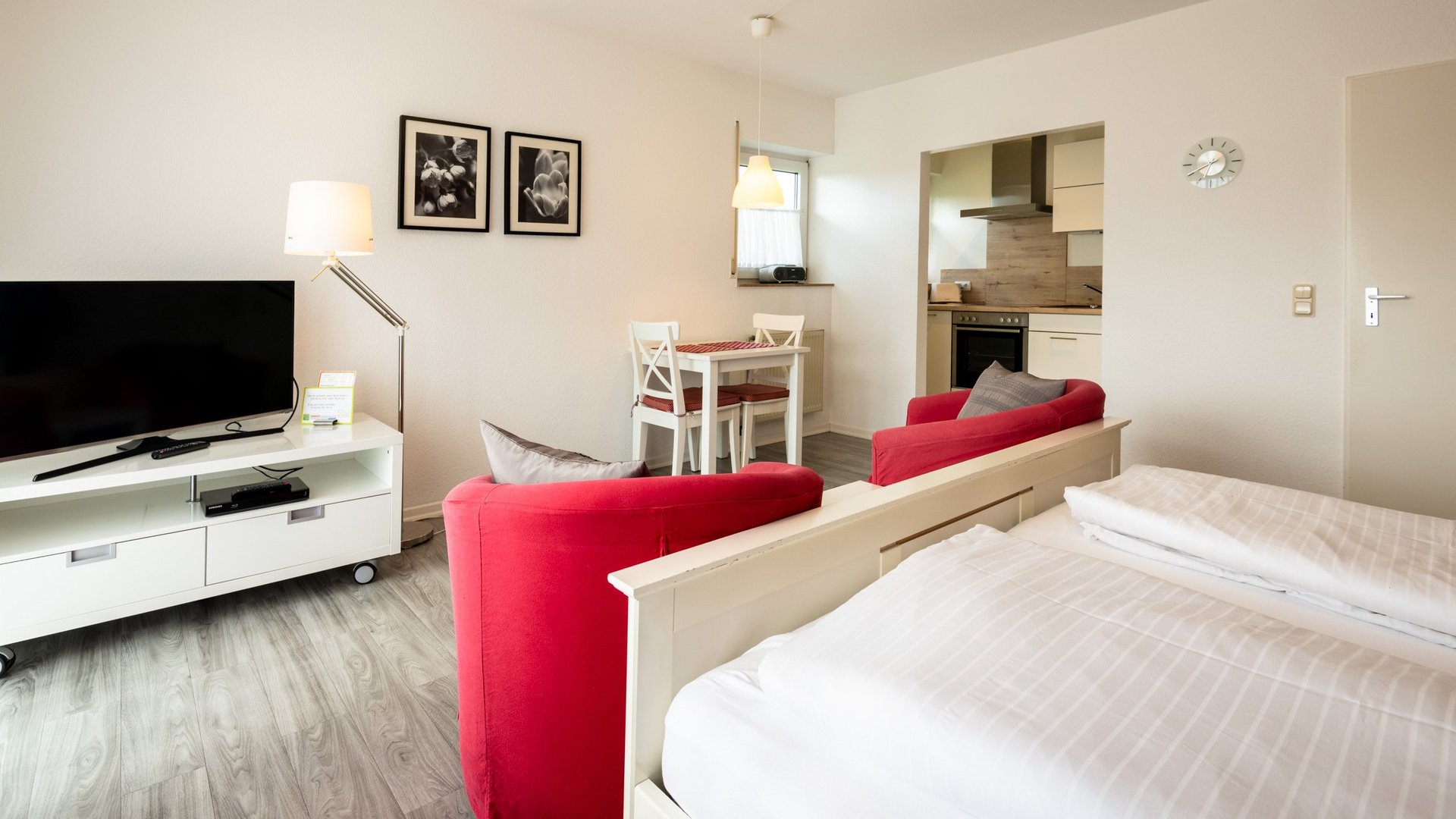 Ihre Unterkunft in Gemünd – Langzeitaufenthalt mit extra Service Helle Einzimmerwohnung mit Bett, roten Sesseln, Fernseher, Esstisch und kleiner Küche