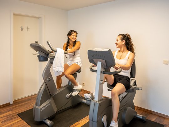 Wohnung mieten in Gemünd? Da haben wir was für Sie! Zwei Frauen unterhalten sich beim Training auf stationären Fahrrädern in einem Raum