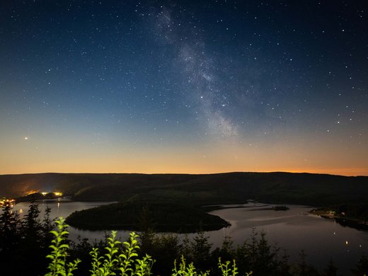 Von der Ferienwohnung Schleiden-Gemünd in den Nationalpark Sternenhimmel mit Milchstraße über einem See und dunklen Hügeln bei Nacht