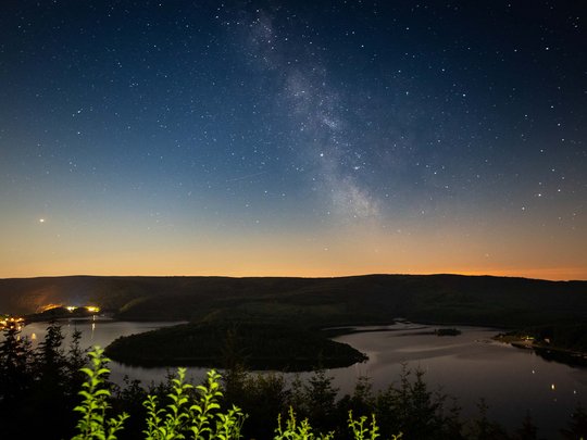 So erreichen Sie unsere Ferienwohnungen in Gemünd. Sternenhimmel mit Milchstraße über einem See und dunklen Hügeln bei Nacht