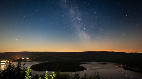 Ferien in der Eifel: ein erster Einblick Sternenhimmel mit Milchstraße über einem See und dunklen Hügeln bei Nacht