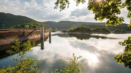 Ferien in der Eifel: ein erster Einblick Staumauer mit Wasser und bewaldeten Hügeln unter bewölktem Himmel