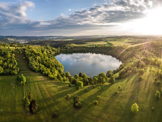 So erreichen Sie unsere Ferienwohnungen in Gemünd. Landschaft mit rundem See, grünen Wiesen und Sonnenstrahlen am Himmel
