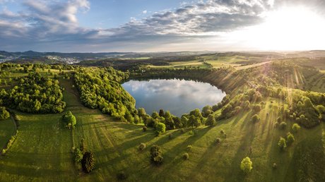Ferien in der Eifel: ein erster Einblick Landschaft mit rundem See, grünen Wiesen und Sonnenstrahlen am Himmel