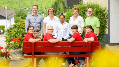 Ferien in der Eifel: ein erster Einblick Gruppe von neun lächelnden Personen, einige auf einer roten Bank sitzend, draußen im Grünen