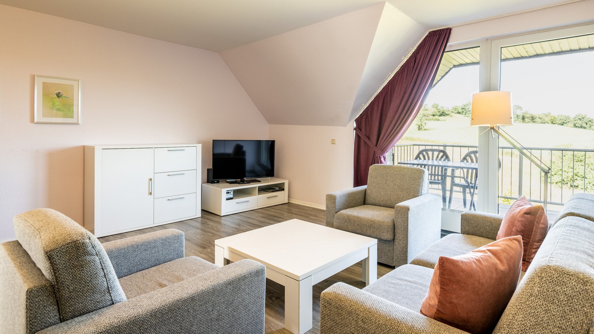 Gemünder Ferienpark: Jobs, die erfüllen Helles Wohnzimmer mit grauen Möbeln, weißem Tisch, TV und Balkon mit Blick ins Grüne