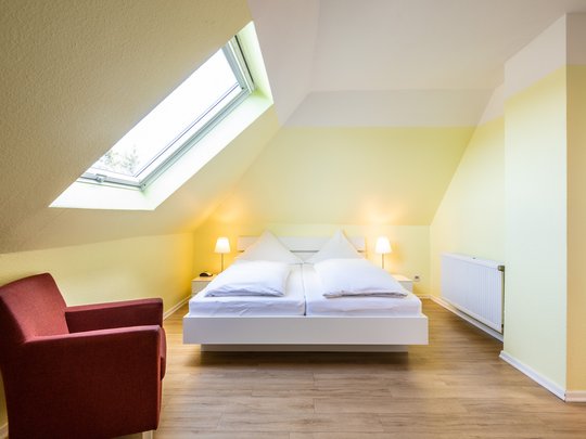 Ihre Unterkunft in Gemünd – Langzeitaufenthalt mit extra Service Helles Schlafzimmer mit Dachfenster, Doppelbett und rotem Sessel