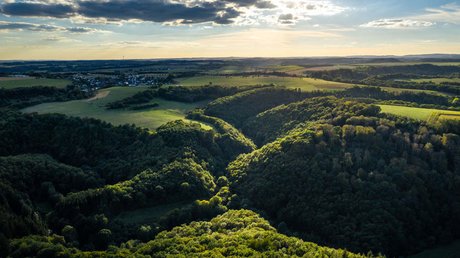 Ferien in der Eifel: ein erster Einblick Luftaufnahme eines bewaldeten Tals mit Feldern und Dorf im Hintergrund bei Sonnenuntergang
