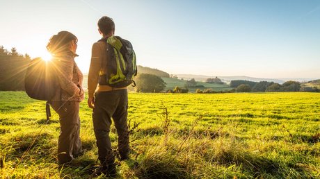 Ferien in der Eifel: ein erster Einblick Zwei Wanderer mit Rucksäcken schauen bei Sonnenuntergang auf eine grüne Landschaft