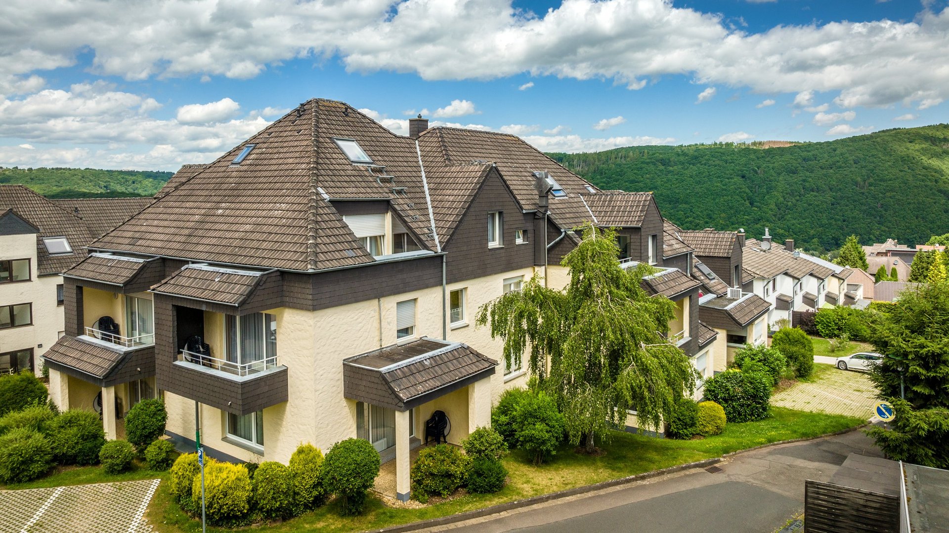 Wohnung mieten in Gemünd? Da haben wir was für Sie! Wohngebäude mit braunen Dächern vor bewaldeten Hügeln und blauem Himmel