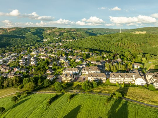 So erreichen Sie unsere Ferienwohnungen in Gemünd. Luftaufnahme eines Dorfes umgeben von grünen Feldern und bewaldeten Hügeln im Sonnenschein