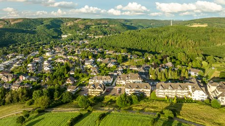 Ferien in der Eifel: ein erster Einblick Luftaufnahme eines Dorfes umgeben von grünen Feldern und bewaldeten Hügeln im Sonnenschein