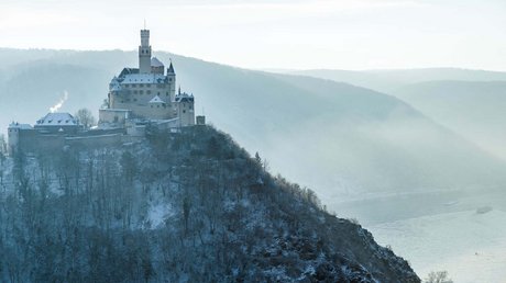 Ferien in der Eifel: ein erster Einblick Schloss auf bewaldetem Hügel im Winter mit Fluss und nebligen Bergen im Hintergrund