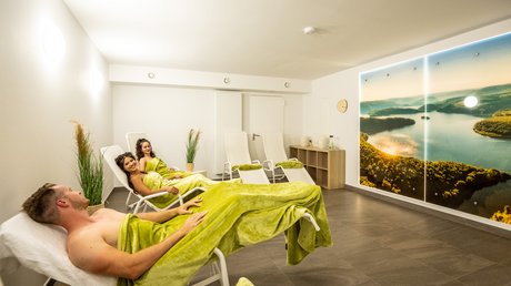 Ferien in der Eifel: ein erster Einblick Drei entspannte Personen in Bademänteln in einem Wellnessruheraum mit Naturbild