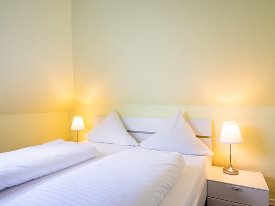 Ihre Unterkunft in Gemünd – Langzeitaufenthalt mit extra Service Gemütliches Schlafzimmer mit weißem Bett, zwei Kissen und zwei Nachttischlampen