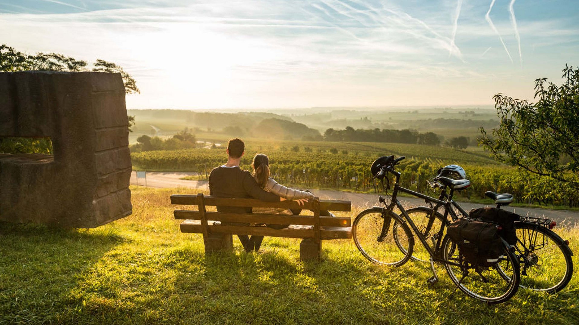 Erlebe die Eifel mit dem Fahrrad – Natur, Freiheit & Komfort Paar sitzt auf Bank mit Fahrrädern und blickt auf sonnige Weinberglandschaft