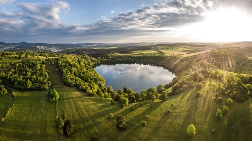Ferienwohnungen in Gemünd mit dem gewissen Extra Landschaft mit rundem See, grünen Wiesen und Sonnenstrahlen am Himmel
