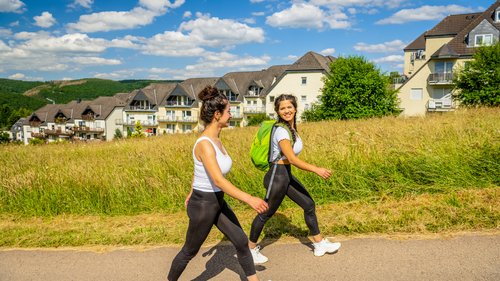 Ferienwohnungen in Gemünd mit dem gewissen Extra Zwei Frauen wandern auf einem Weg mit Häusern und Wiesen im Hintergrund.