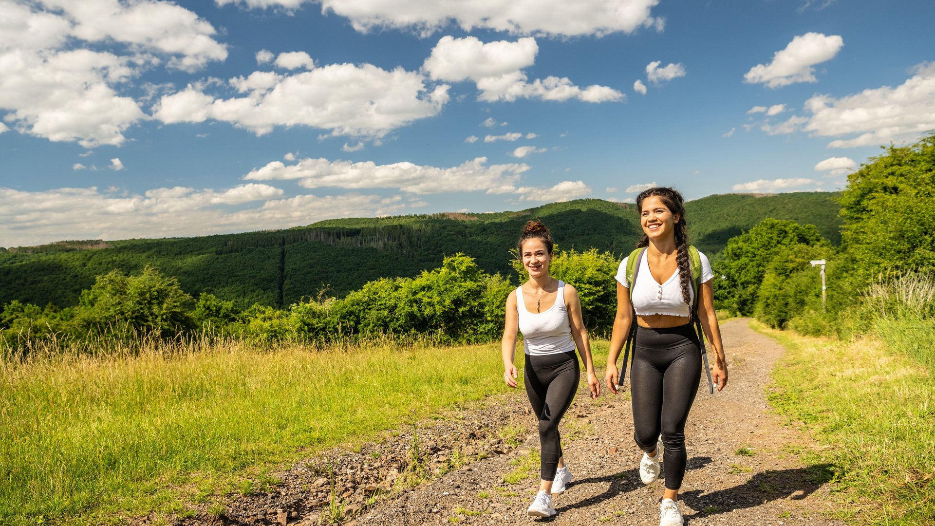 Holidays in the Eifel region: Want a change of pace? Two young women hiking on a trail with green hills and blue sky in the background